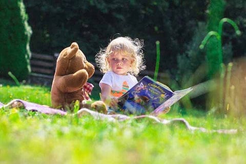 Une petite fille dans un champs entrain de lire un conte pour enfant, accompagnée sa peluche préférée